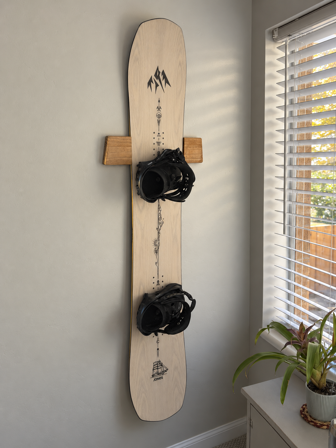 Snowboard with black bindings displayed vertically on a wood wall mount beside a window